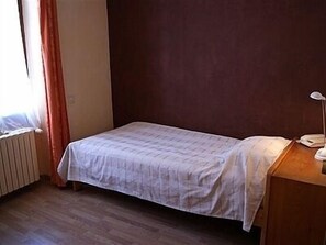 A small, single-bed room with a wooden floor, a bedside table with a lamp, and a window with curtains. - De la Paix (Tournus)