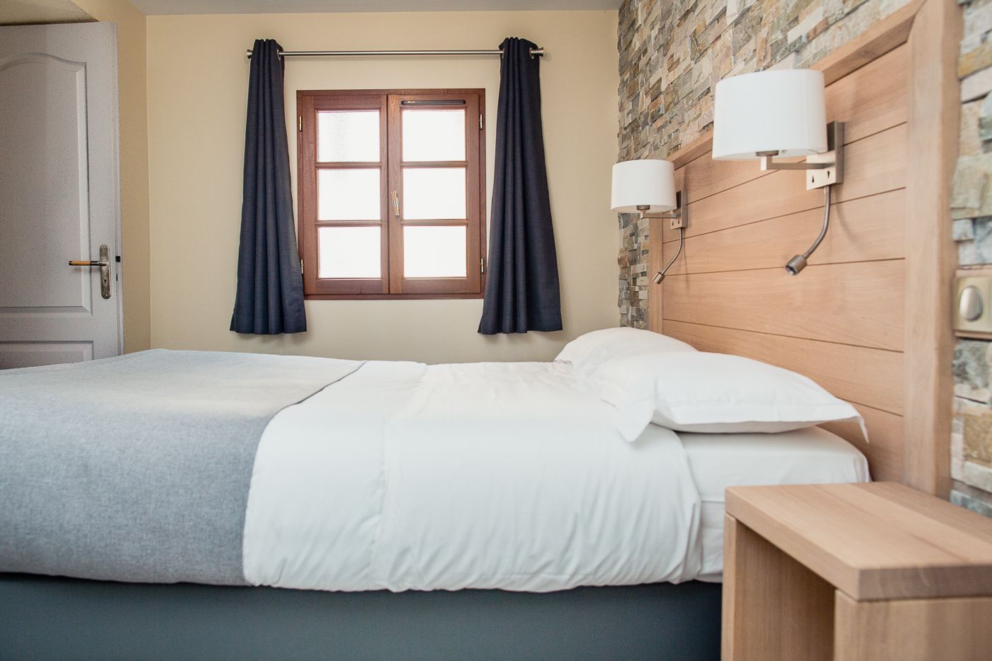 Un lit bien fait, avec du linge de lit blanc, une tête de lit en bois, une table de chevet, une fenêtre avec des rideaux et un mur en pierre.