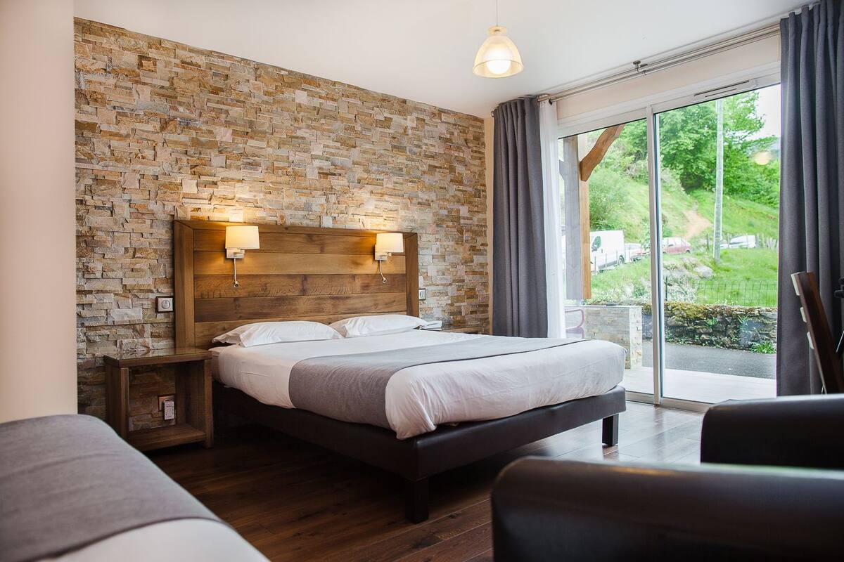 A bedroom with a stone wall, a wooden headboard, a large bed, bedside lamps, a view of greenery through a sliding glass door, and a wooden floor.