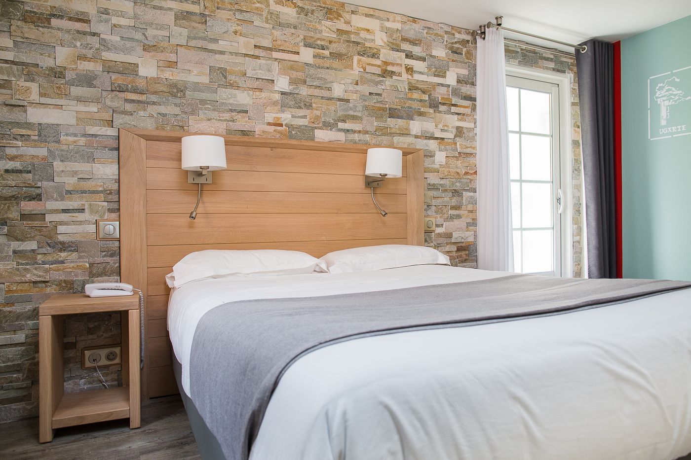 Una cama bien hecha con cabecera de madera, una mesita de noche con lámpara y una pared con diseño de piedra.