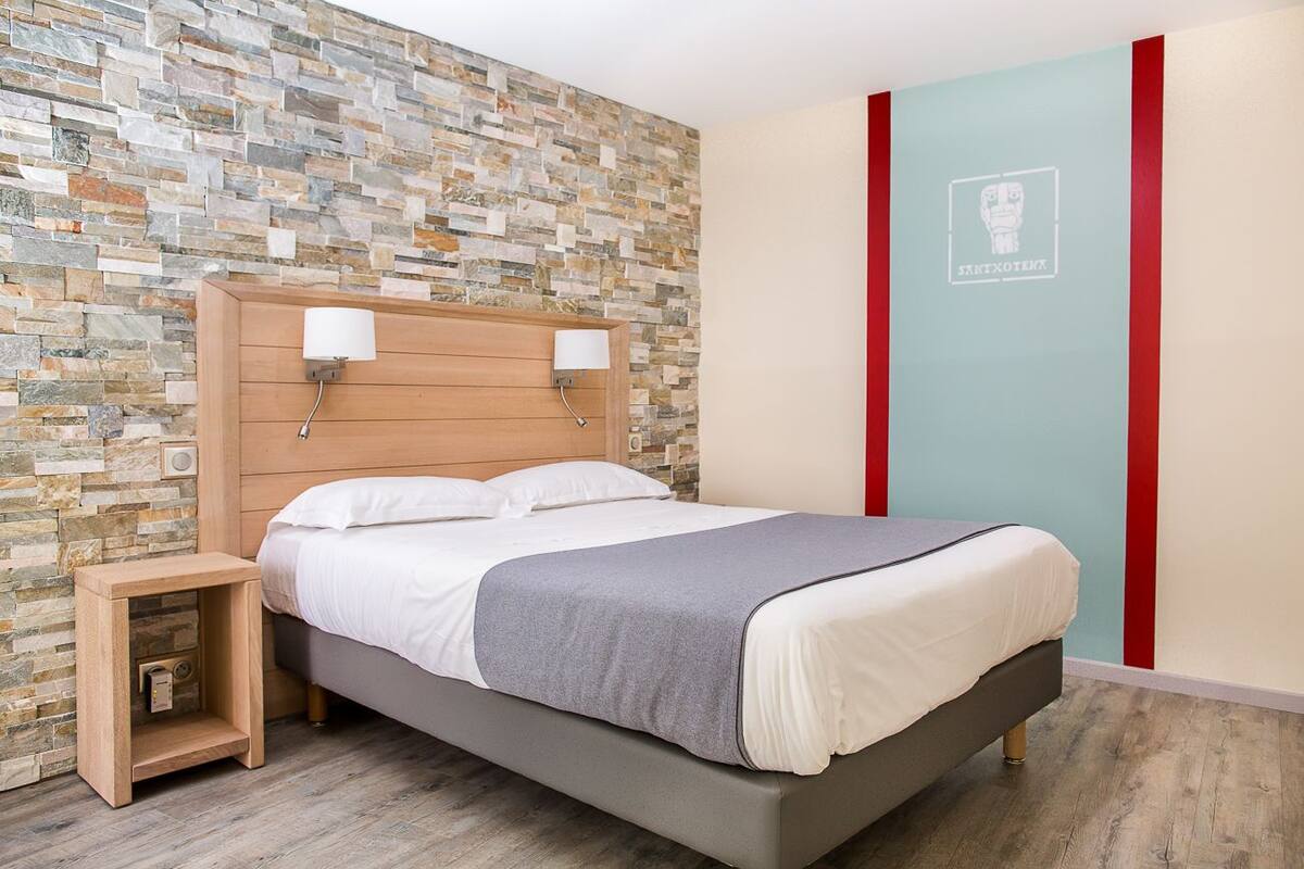A modern bedroom with a stone-patterned wall, a bed with white bedding, a nightstand, and a red and blue accent wall.
