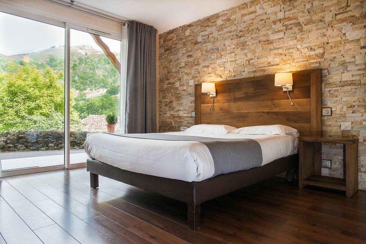 A bedroom with a large bed, wooden headboard, stone wall, and a view of greenery through a sliding glass door.