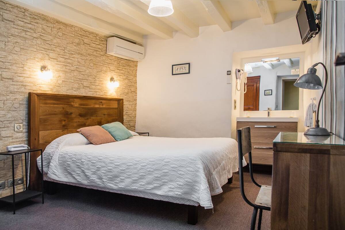 A bedroom with a stone wall, a wooden headboard, a bed with a white quilt, a bedside table, a lamp, a chair, and a desk with a mirror.