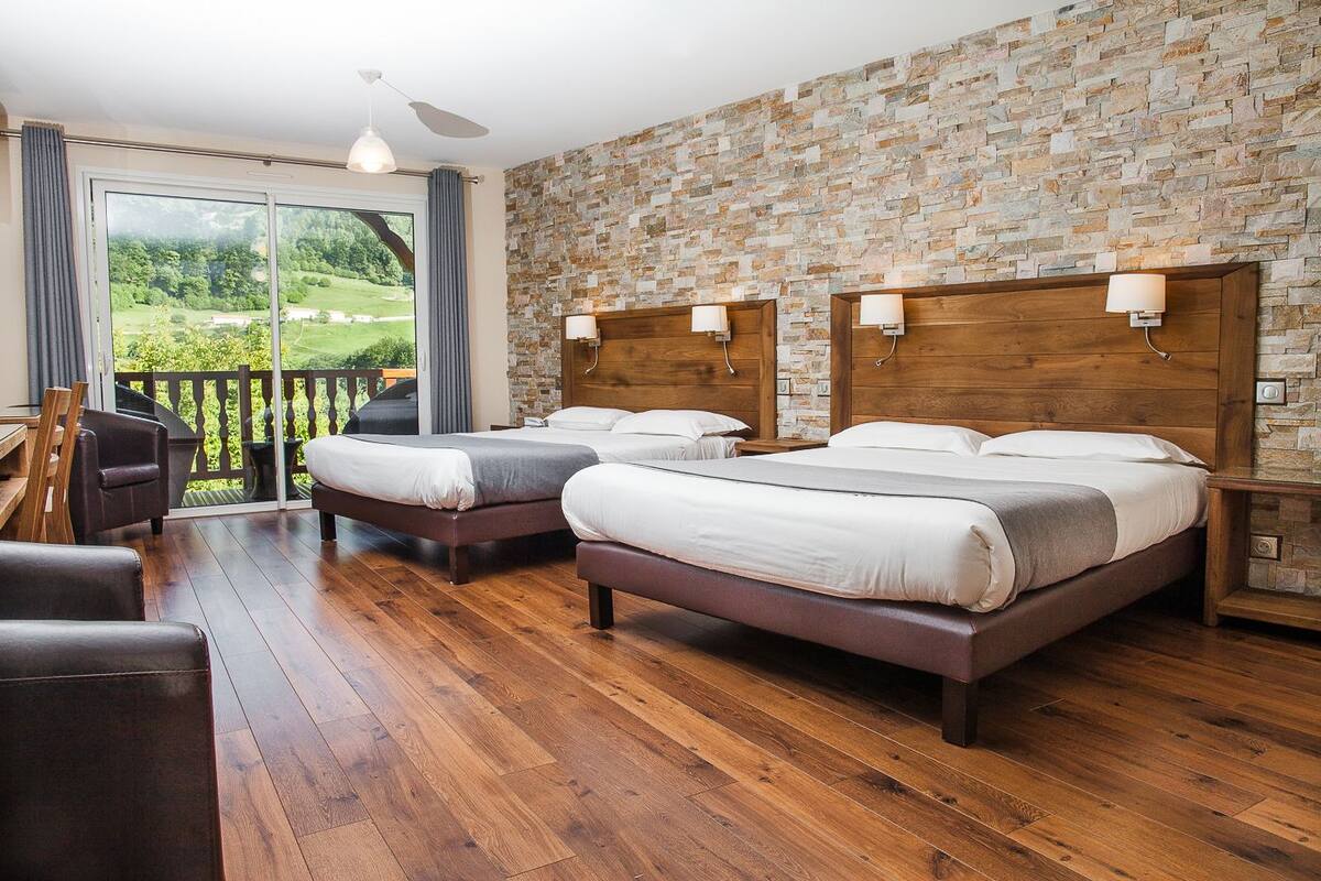 A hotel room with two beds, a wooden floor, a stone wall, and a balcony with a view of greenery.