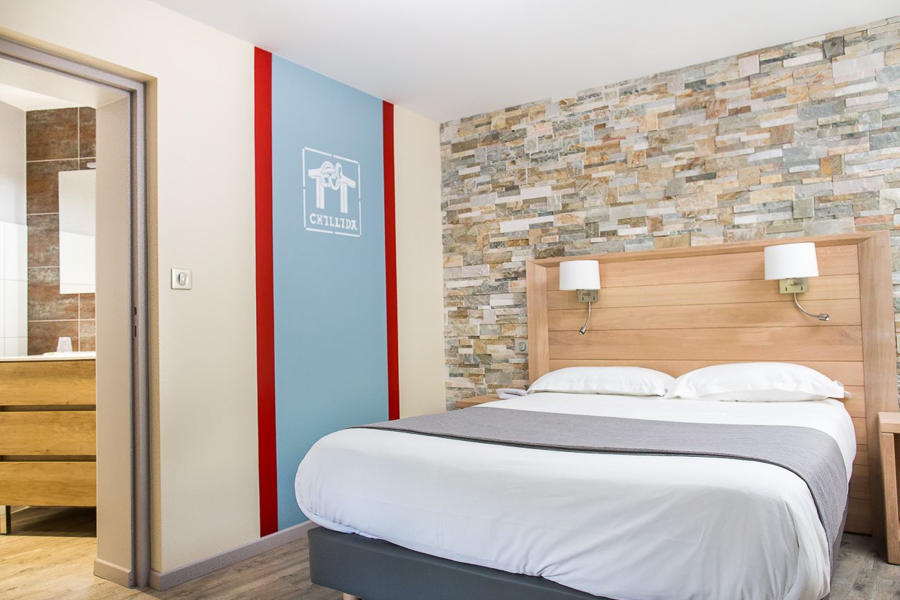 Une chambre d’hôtel avec un lit, une tête de lit en bois, un mur en pierre et une enseigne indiquant le nom de l’hôtel.