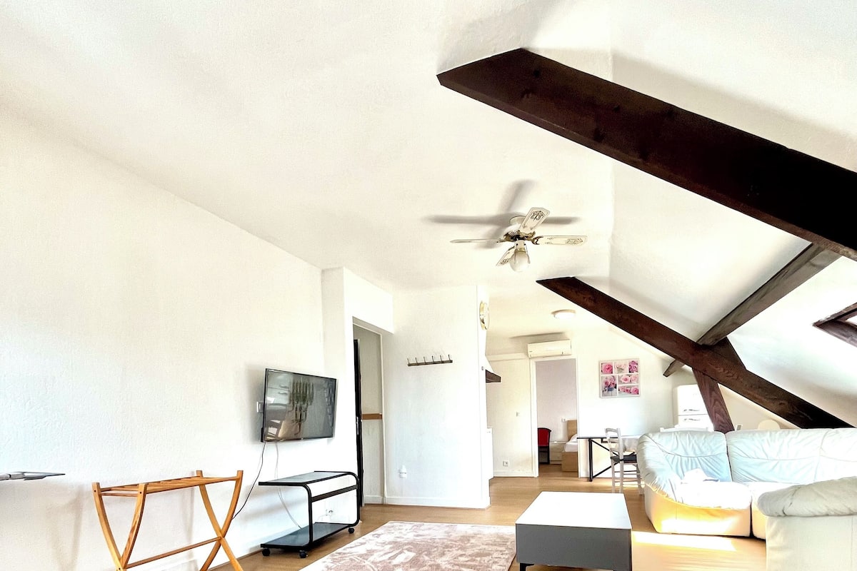 A spacious loft bedroom with a sloped ceiling, a ceiling fan, a large window, and a view of the city.