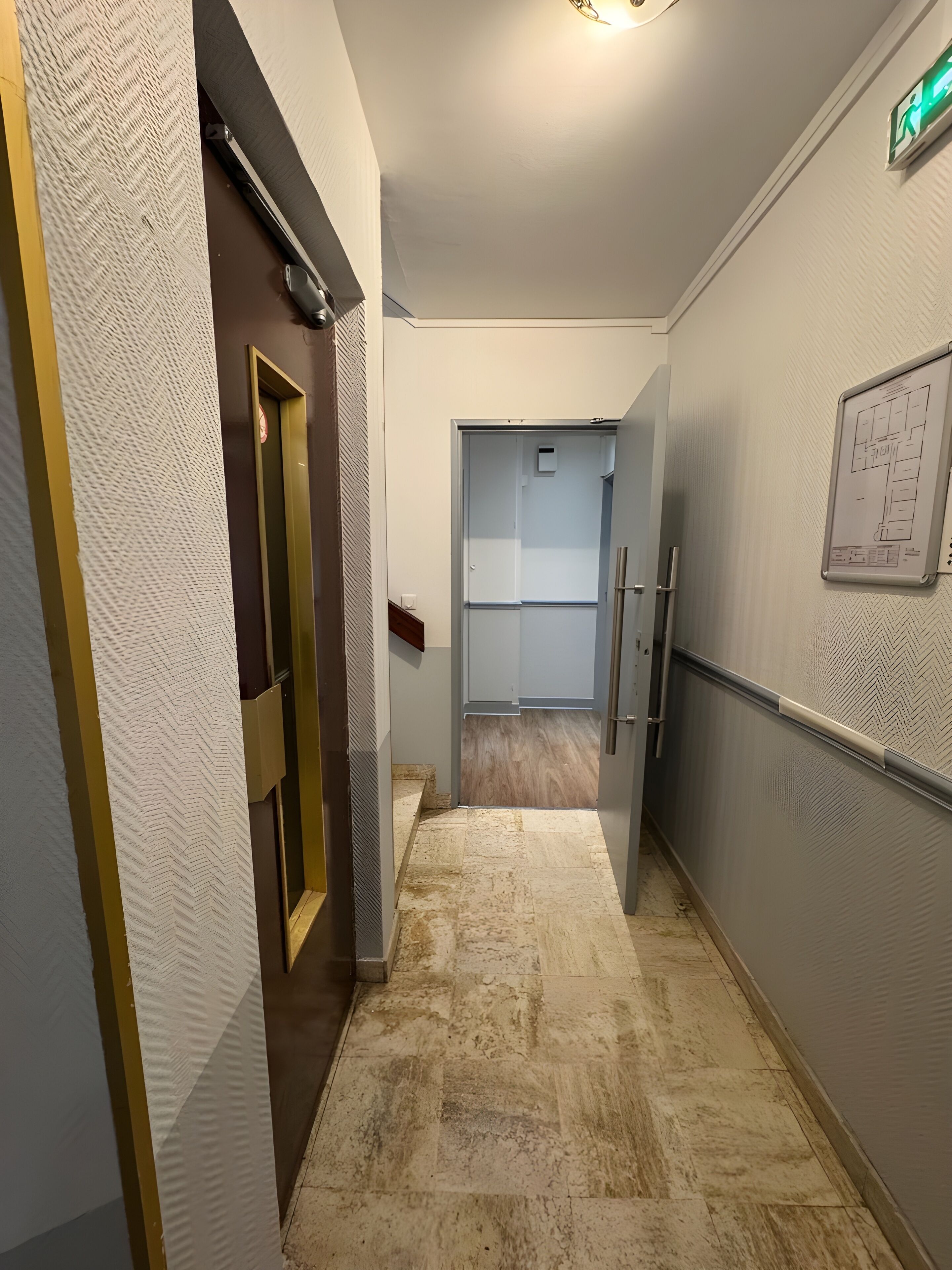 Un couloir étroit, avec un mur texturé sur le côté gauche, une porte dotée d’un cadre doré et un sol carrelé.