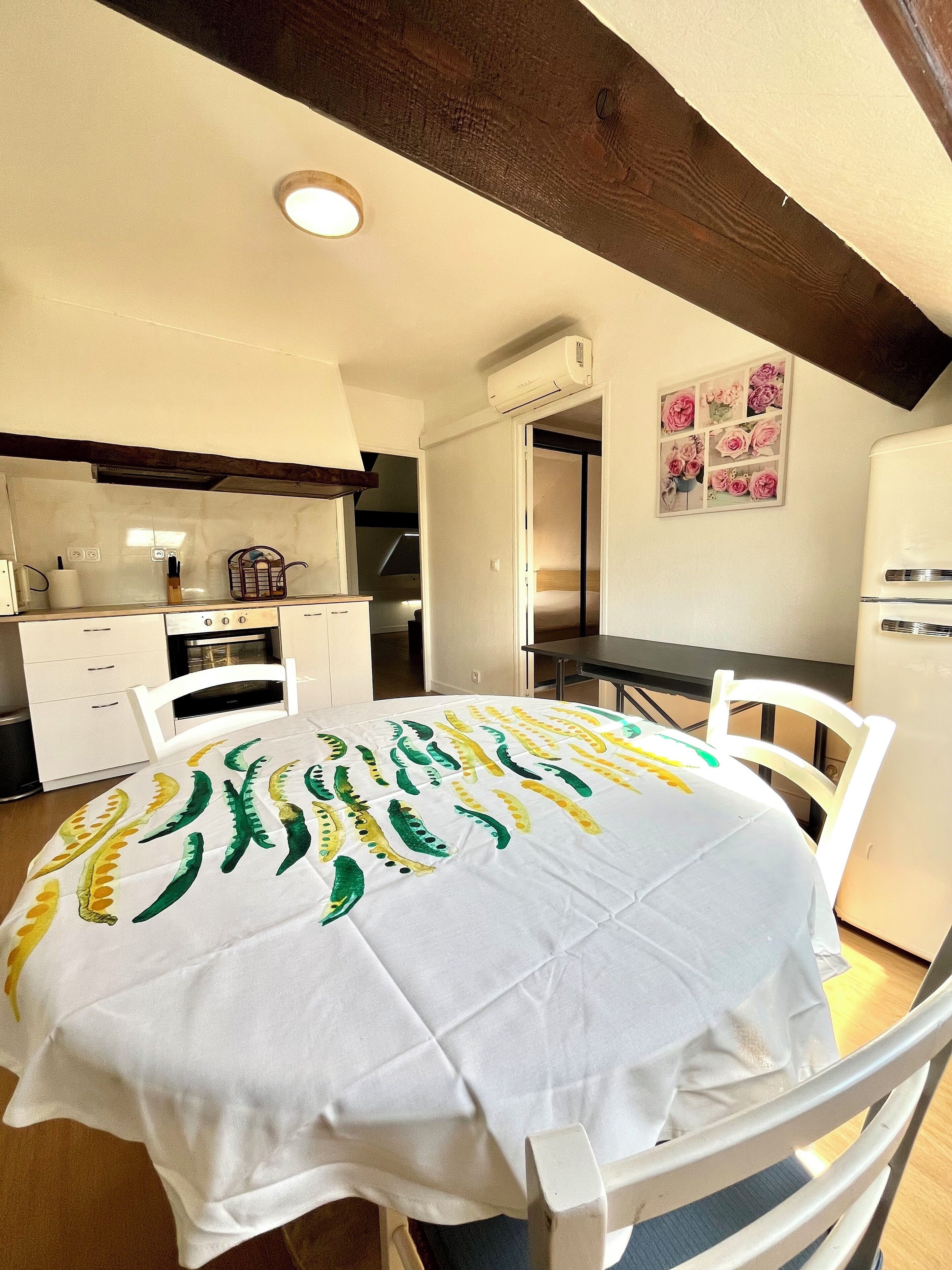 Un coin repas avec une table recouverte d’une nappe blanche ornée de motifs verts et jaunes, entourée de chaises blanches.