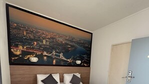 A neatly made bed with white linens and a gray throw, a large cityscape painting above it, and a wooden floor. - Hôtel du Parc (Aulnay-sous-Bois)