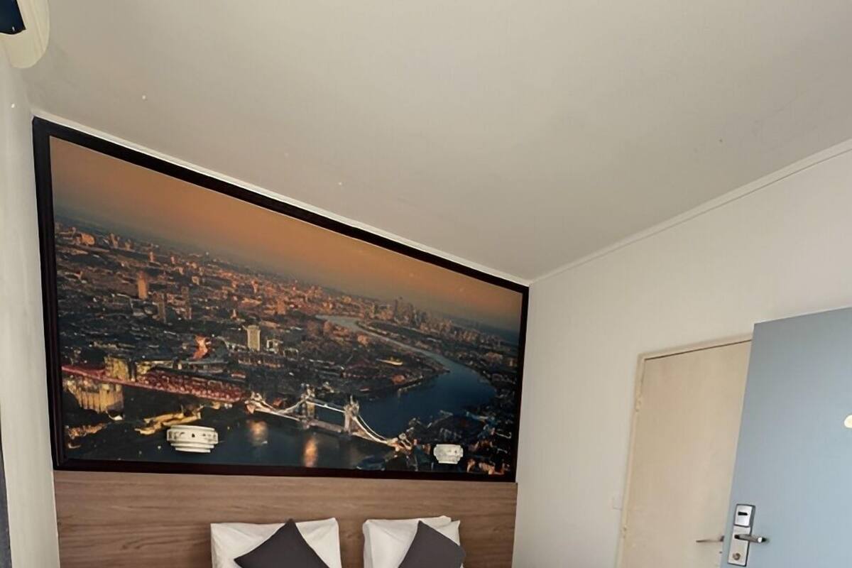 A neatly made bed with white linens and a gray throw, a large cityscape painting above it, and a wooden floor.