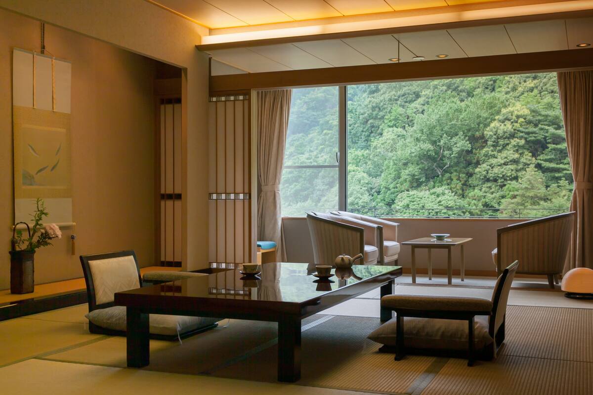 Een traditionele Japanse kamer met tatami vloerbedekking, een lage tafel en uitzicht op een bos door een groot raam.