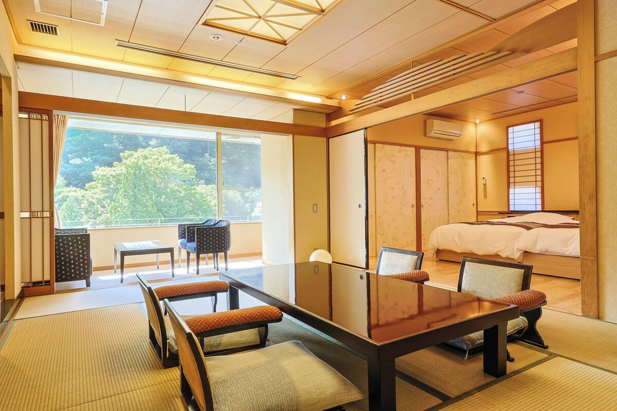 Een traditionele Japanse kamer met tatami vloerbedekking, een lage tafel met kussens, een bed met houten hoofdeinde en grote ramen met uitzicht op groen.