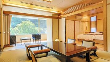 A traditional Japanese-style room with tatami flooring, a low table with cushions, a bed with a wooden headboard, and large windows offering a view of greenery.