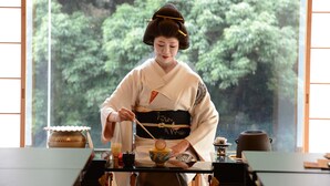 Une scène typique du Japon traditionnel, avec une personne vêtue d’un kimono, une table sur laquelle se trouve une théière, et une vue sur la verdure à travers une fenêtre.