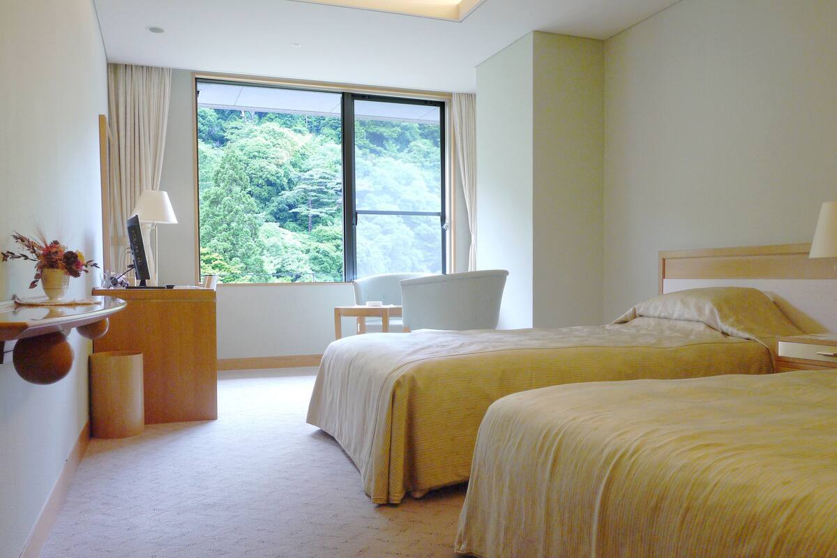 Een hotelkamer met twee bedden, een bureau met een lamp, een stoel en uitzicht op groen door het raam.