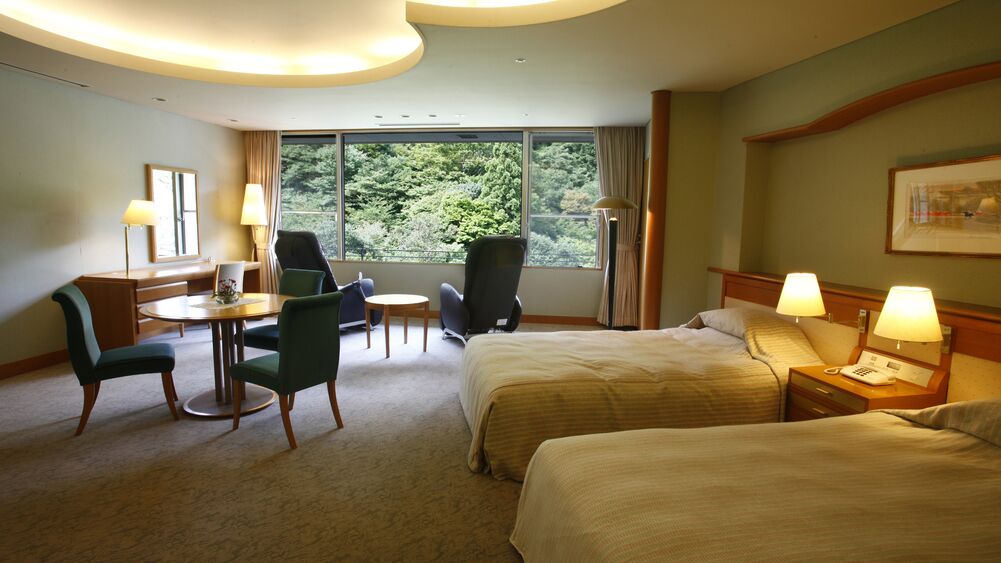 A hotel room with two beds, a desk with a chair, a small table, and a view of greenery through the window.