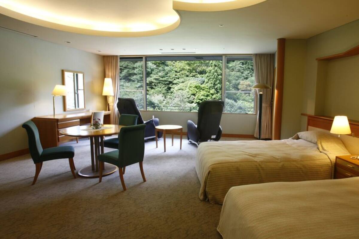 Een hotelkamer met twee bedden, een bureau met stoel, een kleine tafel en uitzicht op groen door het raam.