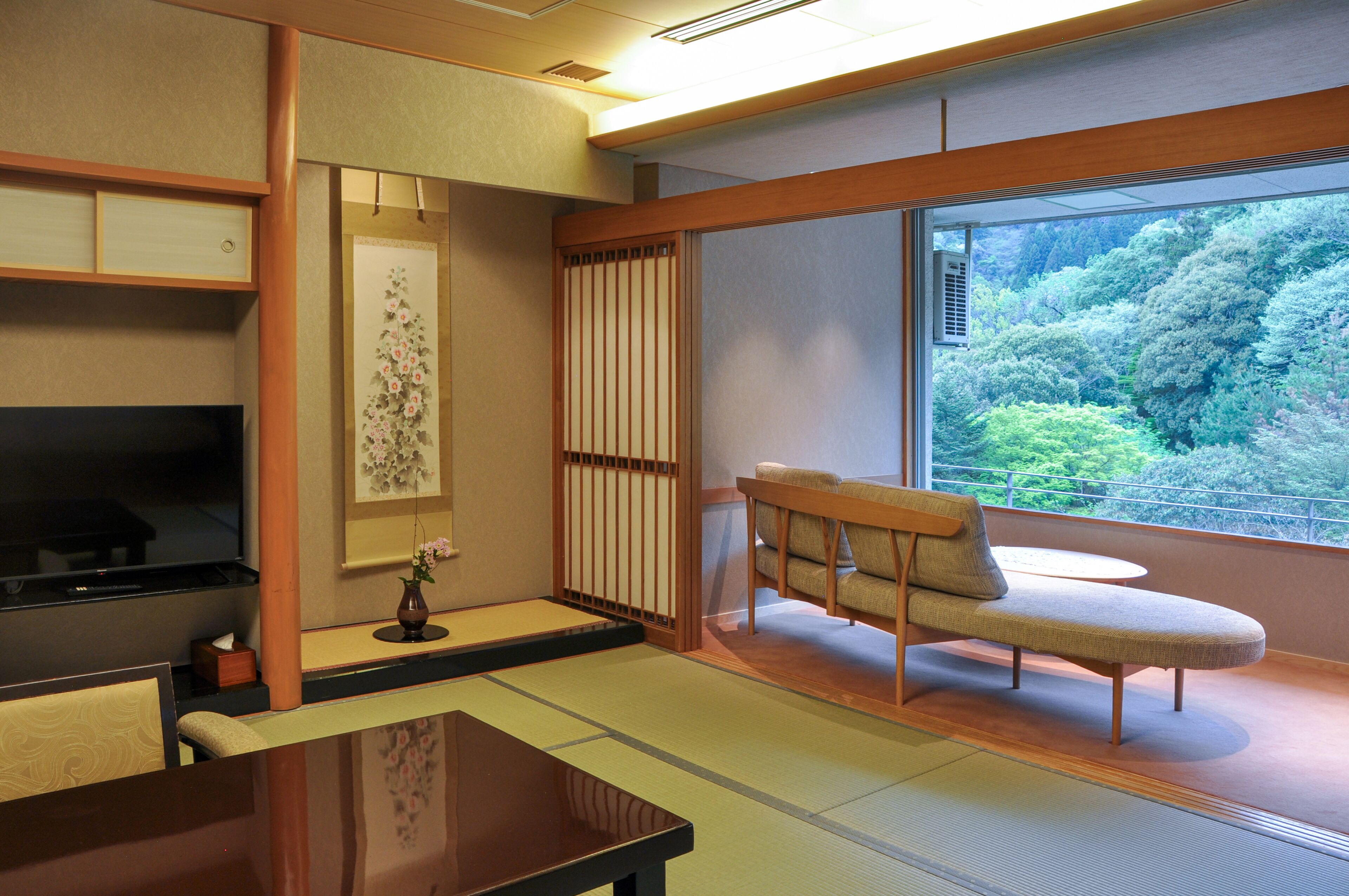 A traditional Japanese room with tatami flooring, a low table, a sofa, and a sliding door leading to a balcony with a forest view.