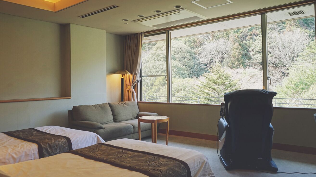 A hotel room with two beds, a sofa, a small table, a massage chair, and a large window with a forest view.