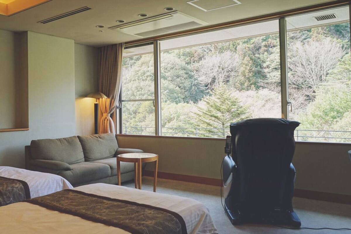 Een hotelkamer met twee bedden, een bank, een kleine tafel, een massagestoel en een groot raam met uitzicht op een bos.