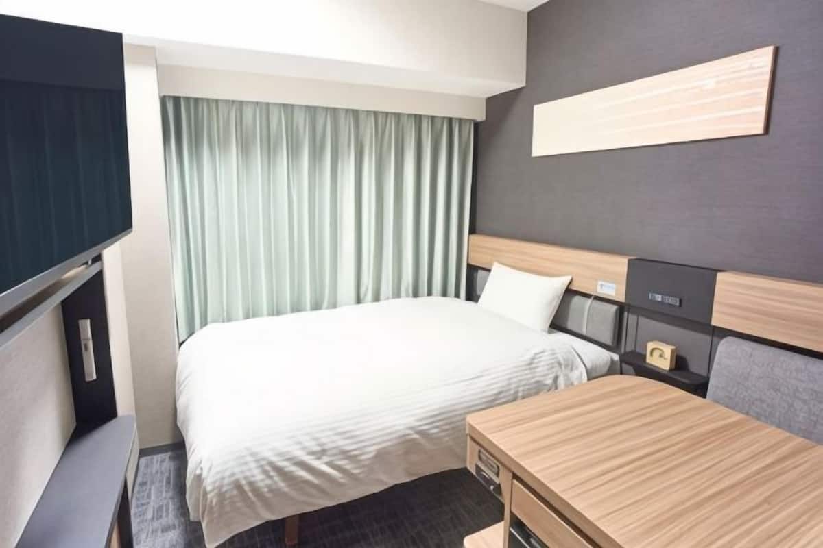 A hotel room with a bed, bedside table, TV, and a view of the outside.