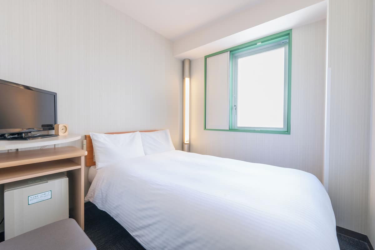 A hotel room with a bed, a desk with a television, a small refrigerator, and a window with a green frame.