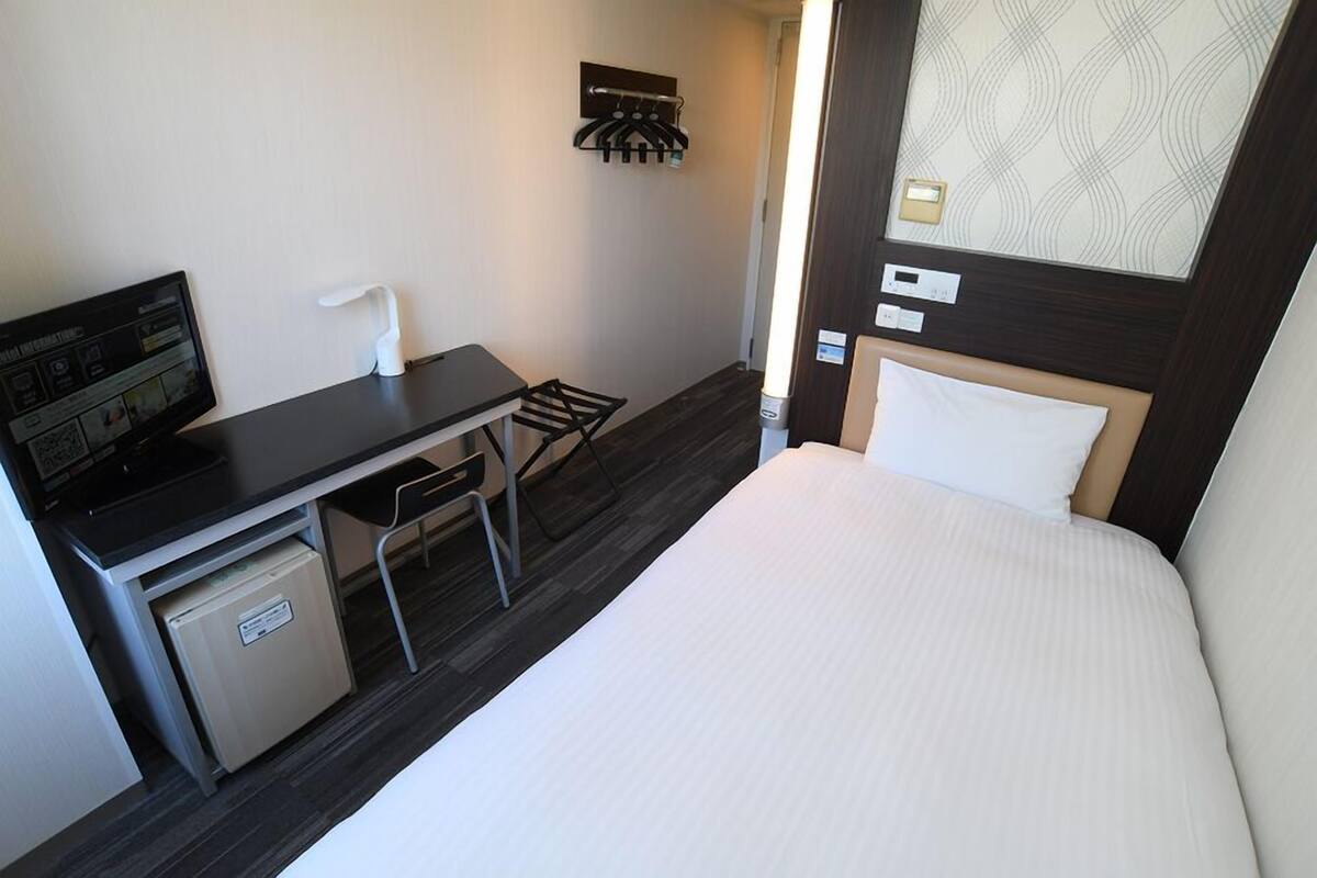 Une chambre d’hôtel équipée d’un lit, d’un bureau avec un ordinateur, d’un petit réfrigérateur et d’un porte-serviettes fixé au mur.