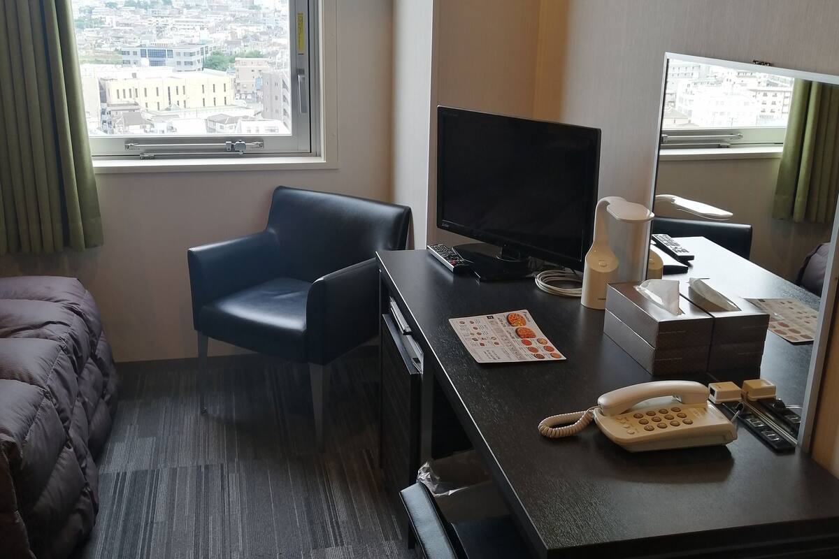 Une chambre d’hôtel avec un lit, un bureau équipé d’un ordinateur, un téléphone et une fenêtre donnant sur la ville.