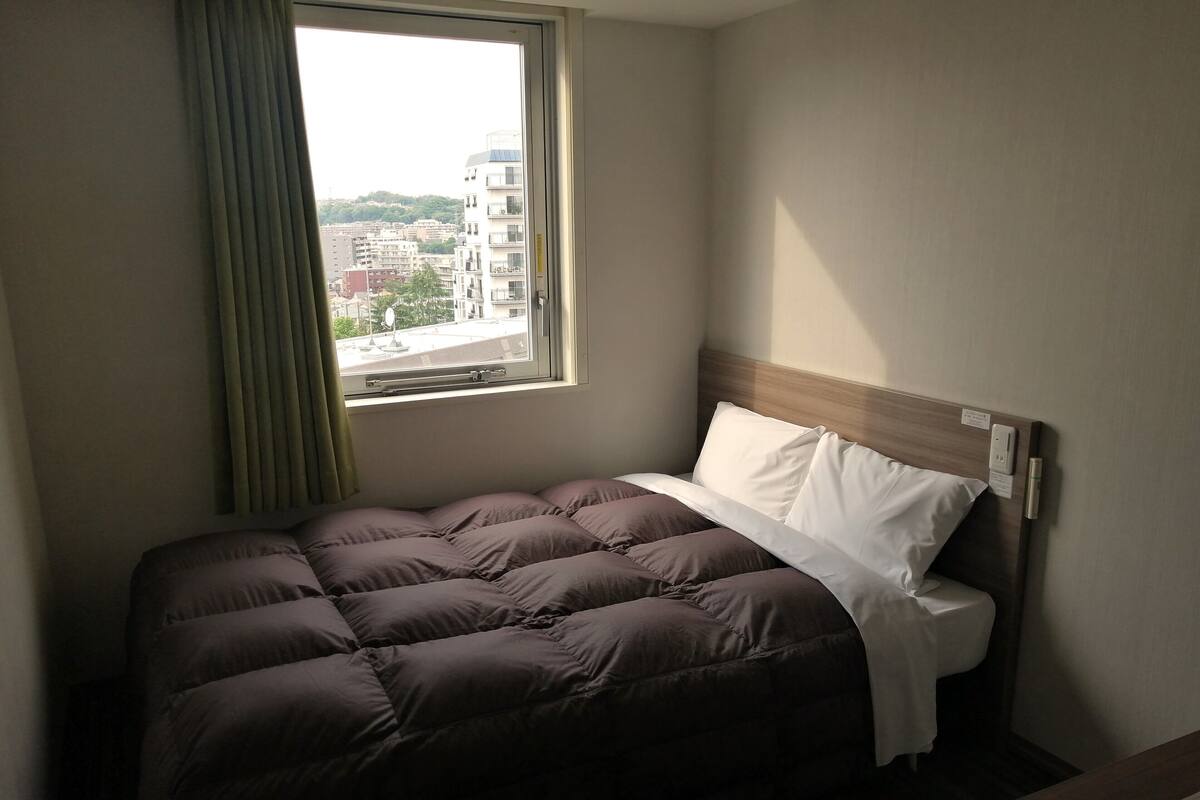 Un lit bien fait, avec une tête de lit, une fenêtre avec des rideaux et une vue sur les bâtiments à l’extérieur.