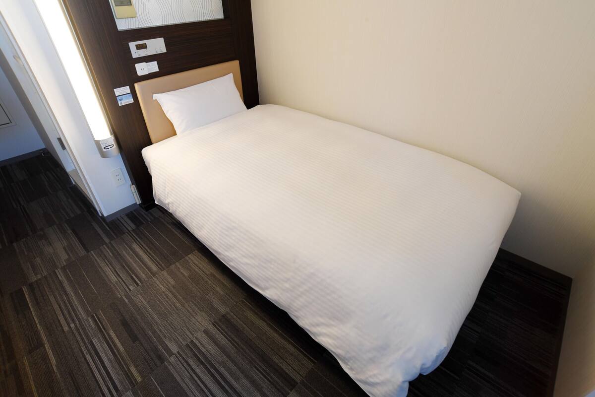 Un lit simple avec un matelas et un oreiller blancs, dans une pièce dont le sol est recouvert de parquet foncé et qui est agrémentée d’une tête de lit en bois.