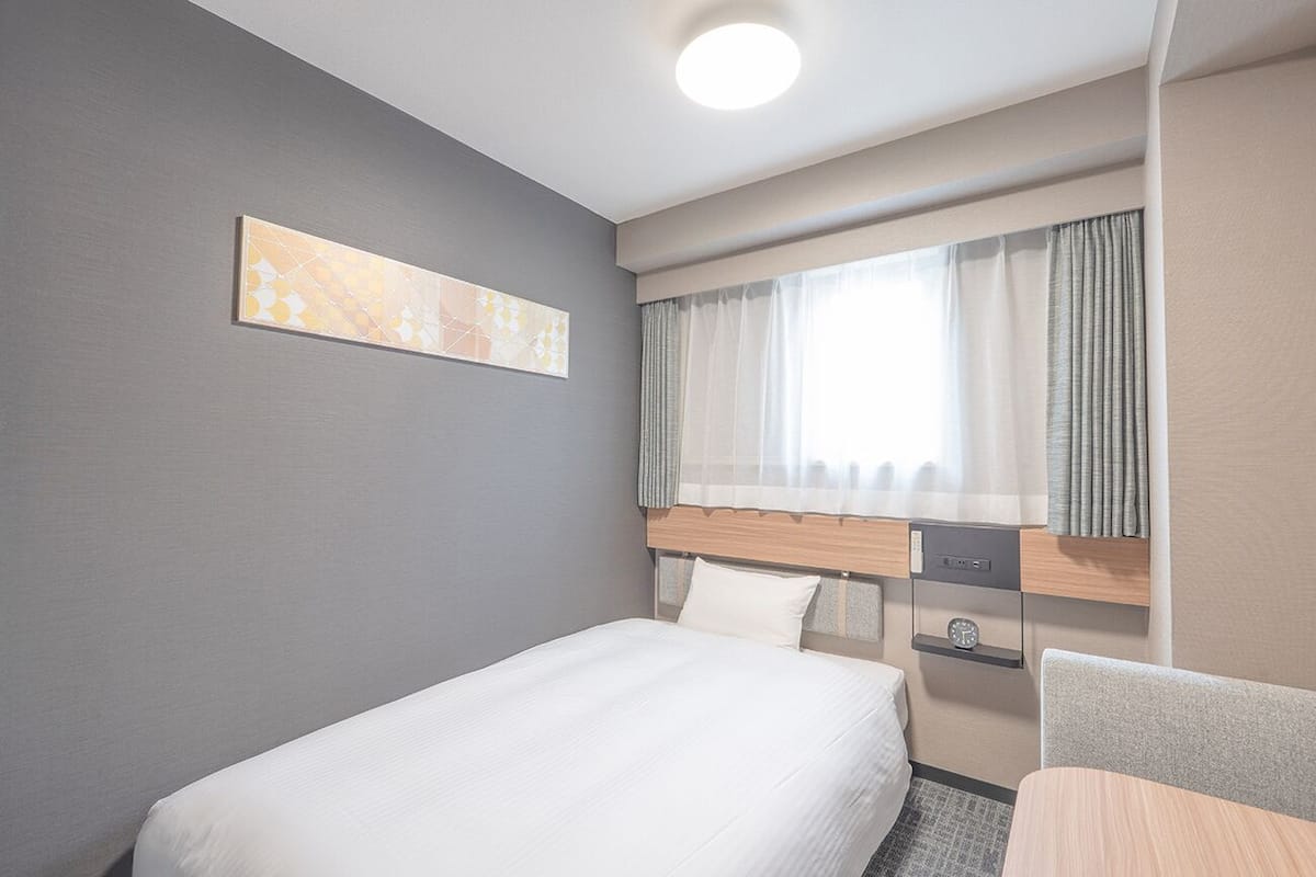Ein Hotelzimmer mit einem Einzelbett, einem kleinen Tisch, einem Stuhl, einem Fenster mit Vorhängen, einem Bild an der Wand und einer Uhr auf dem Nachttisch.