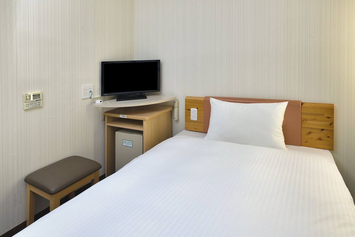 A hotel room with a bed, a television on a desk, a small cabinet, and a bench.