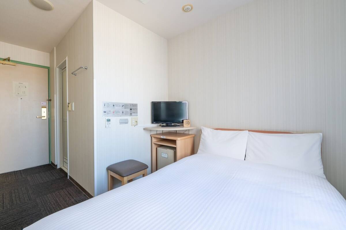 Habitación de hotel con una cama, un televisor sobre un atril de madera, un taburete pequeño y una puerta con marco verde.