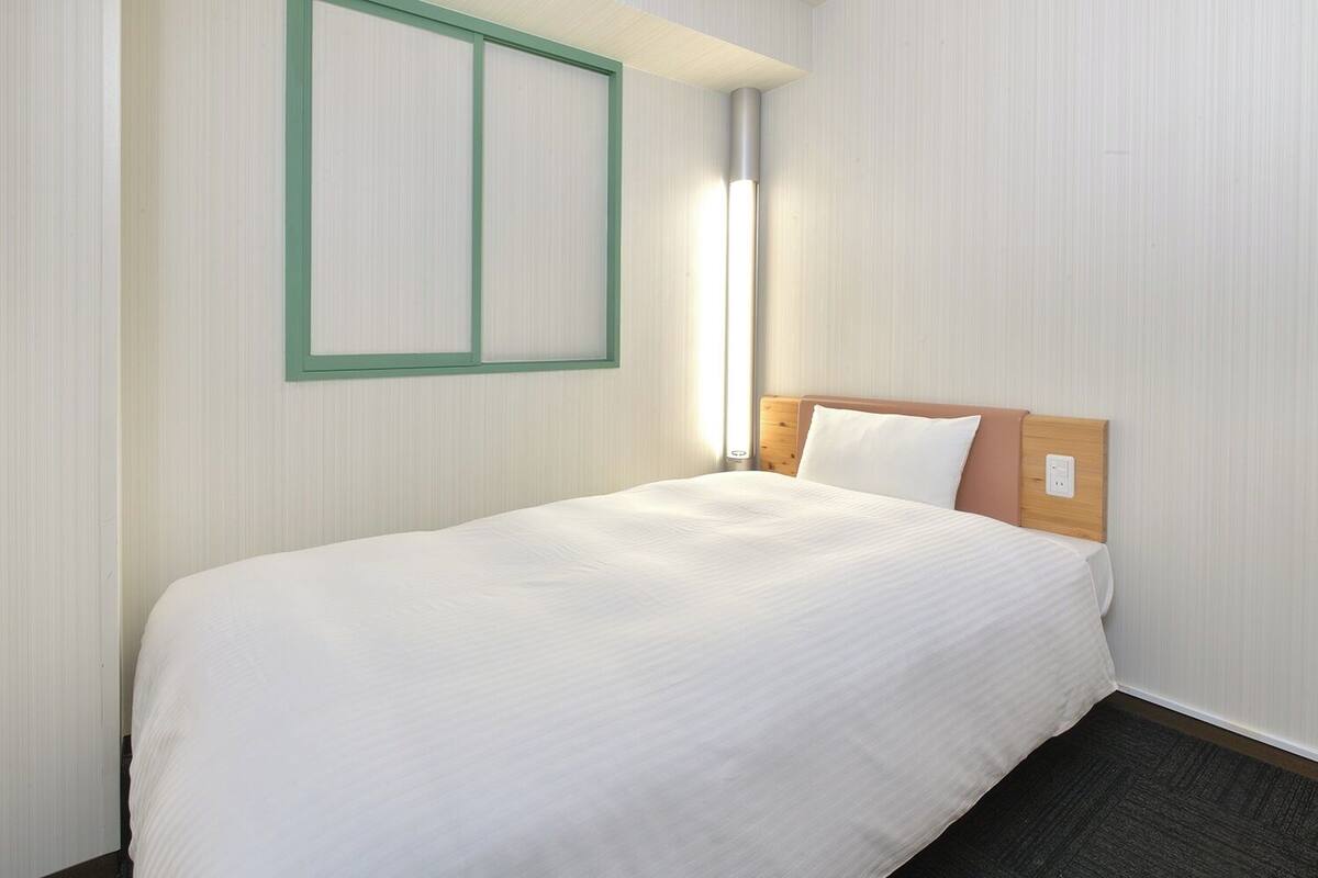 Habitación de hotel con una cama individual, cabecero de madera, ventana con marco verde y una lámpara fijada a la pared.