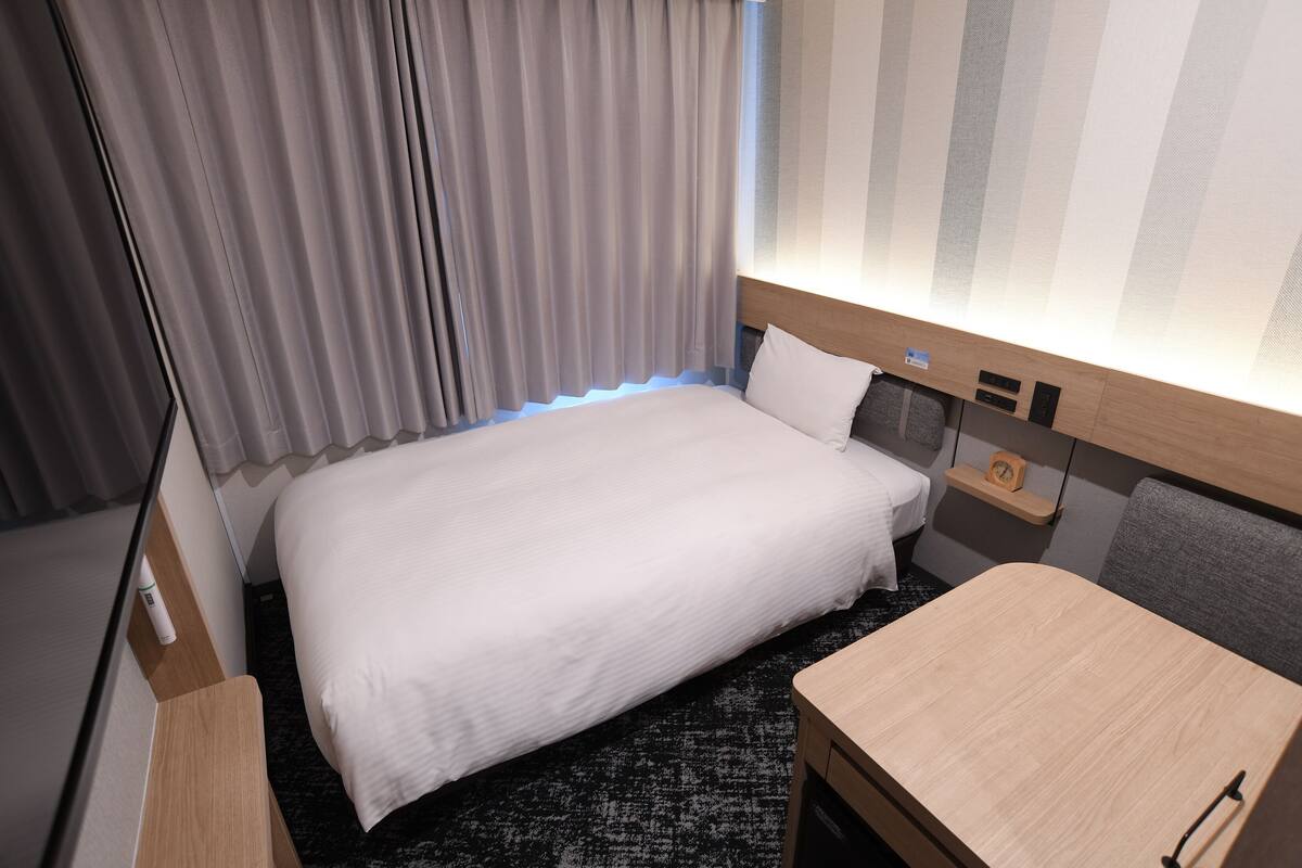Una habitación de hotel con una cama bien hecha, una mesita de noche, un pequeño estante con un reloj y un cabecero con iluminación integrada.