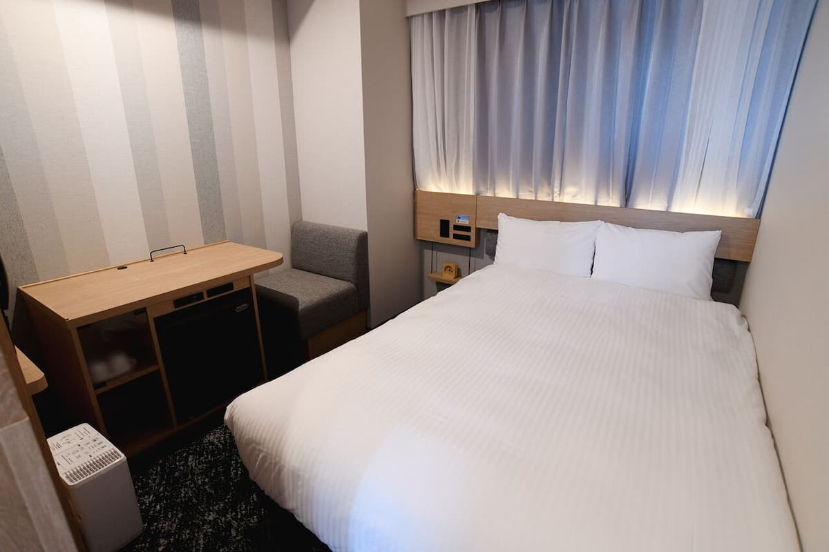 Una habitación de hotel con cama, escritorio, silla, mesita pequeña y mesita de noche.