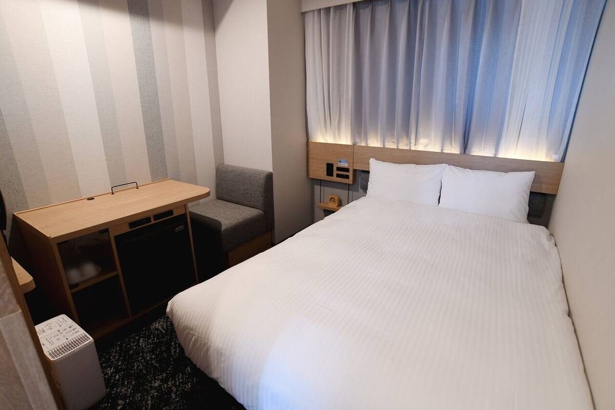A hotel room with a bed, a desk, a chair, a small table, and a bedside table.