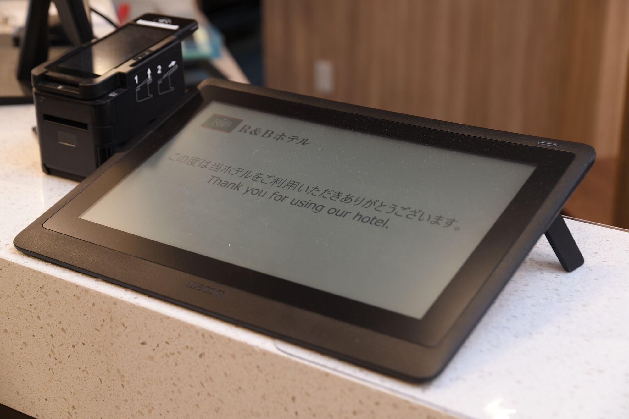 Une tablette affiche un message de remerciement en anglais et en japonais. Elle est posée sur un comptoir, à côté d’une station de recharge noire, avec un mur blanc en arrière-plan.