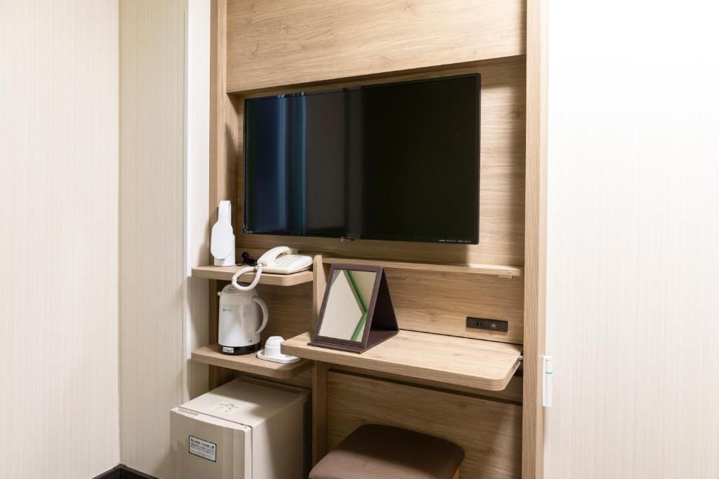 Habitación de hotel funcional con cama, un escritorio pequeño con teléfono y cafetera, un televisor de pantalla plana en la pared y un mueble auxiliar.