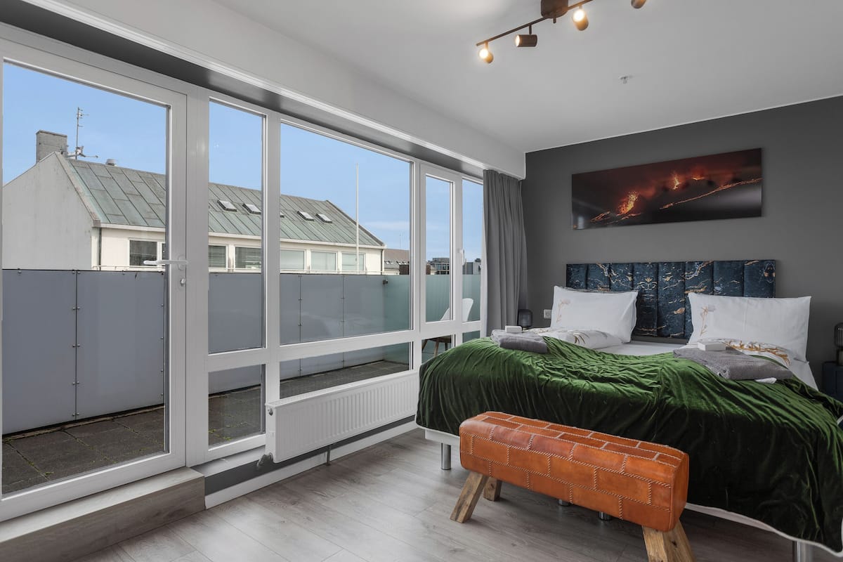A bedroom with a large window, a bed with green bedding, a bench, and a painting on the wall.