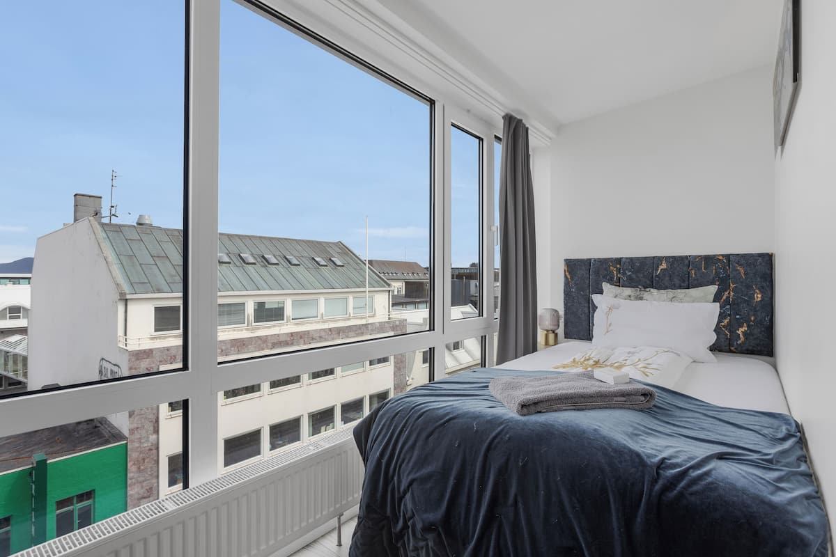 A bedroom with a large window, a bed with a dark blue bedspread, and a view of buildings outside.