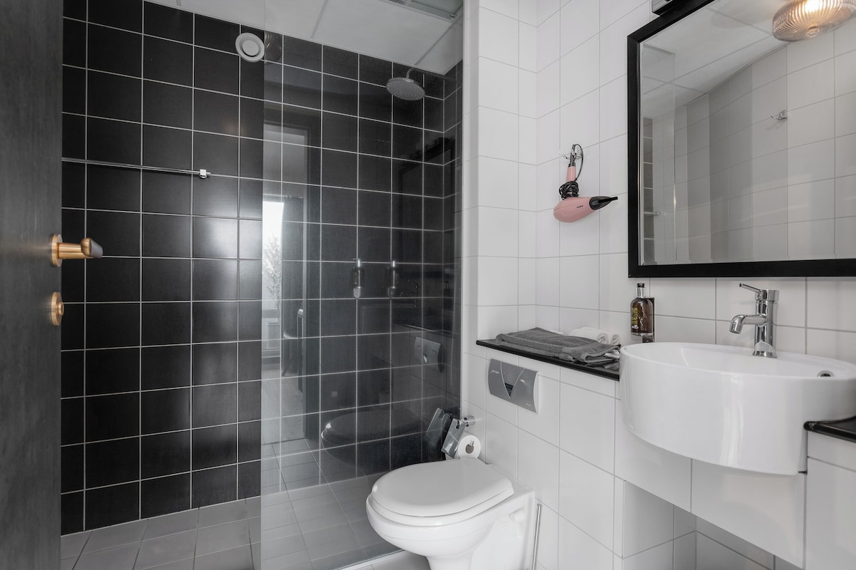 Une salle de bain moderne avec une douche carrelée de noir, des toilettes blanches et un grand miroir au-dessus d’un lavabo blanc.