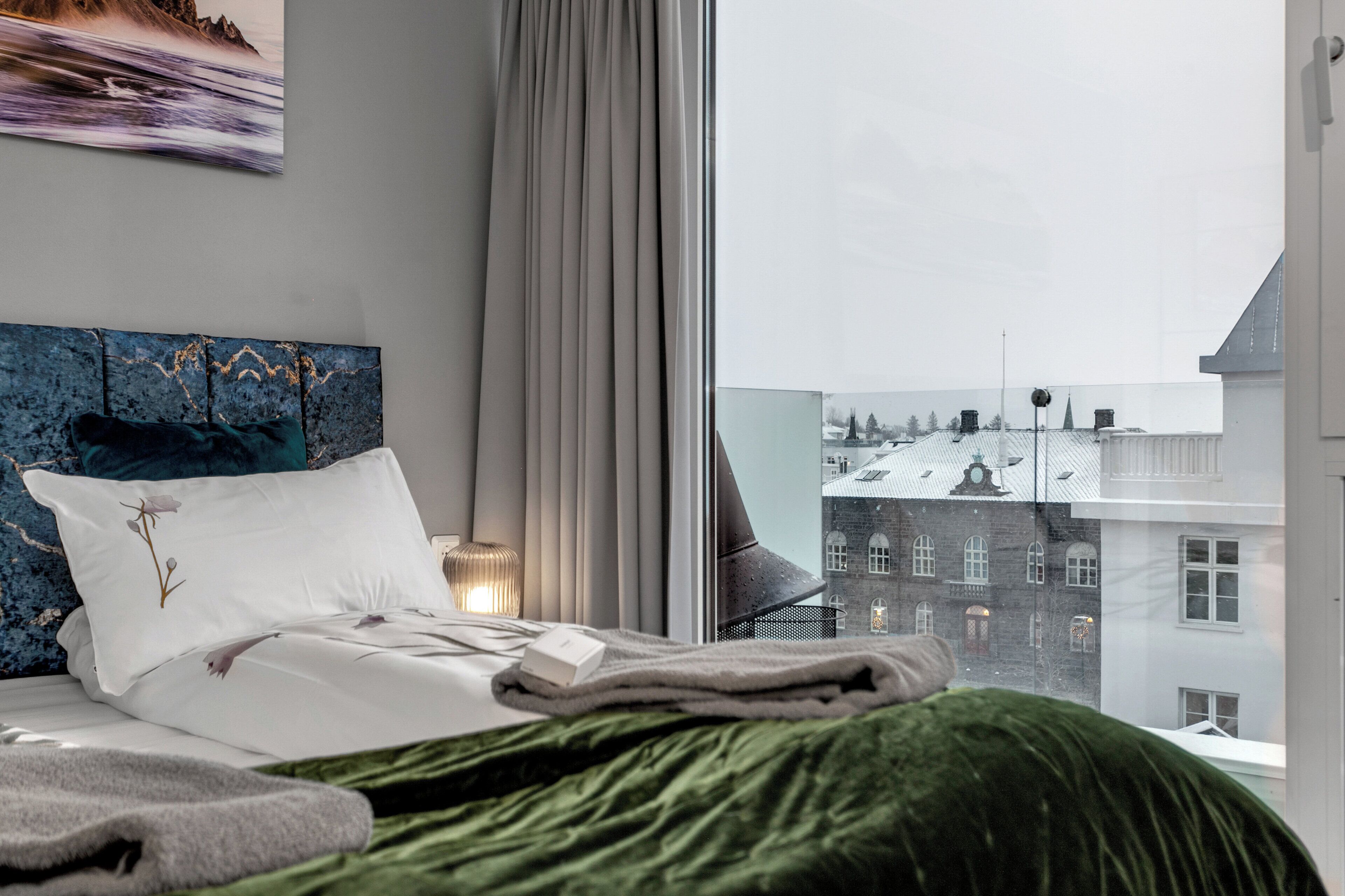 A hotel room with a bed, a view of buildings, and a window with curtains.