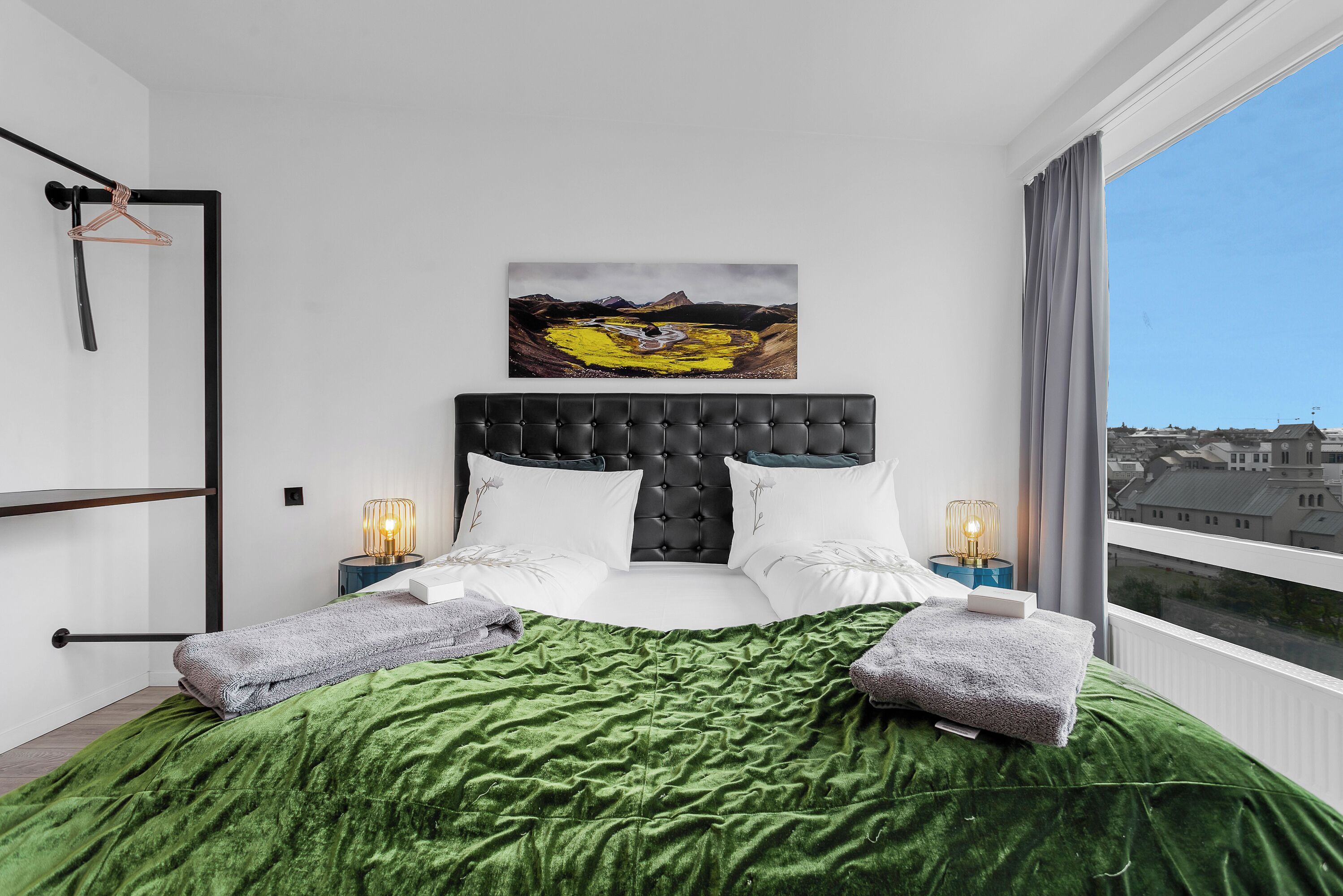 Un dormitorio moderno con colcha de terciopelo verde, cabecera tapizada negra, almohadas blancas, dos lámparas de noche y vistas a la ciudad a través de un ventanal grande.