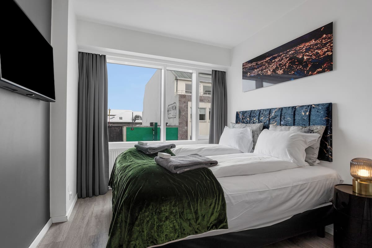 A modern hotel room with a large bed, a flat-screen TV, a cityscape painting, and a view of buildings through the window.