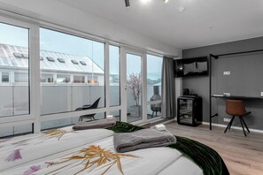 A modern hotel room with a large bed, a desk, a TV, and a view of the cityscape through large windows. - City Center Hotel (Reykjavik)