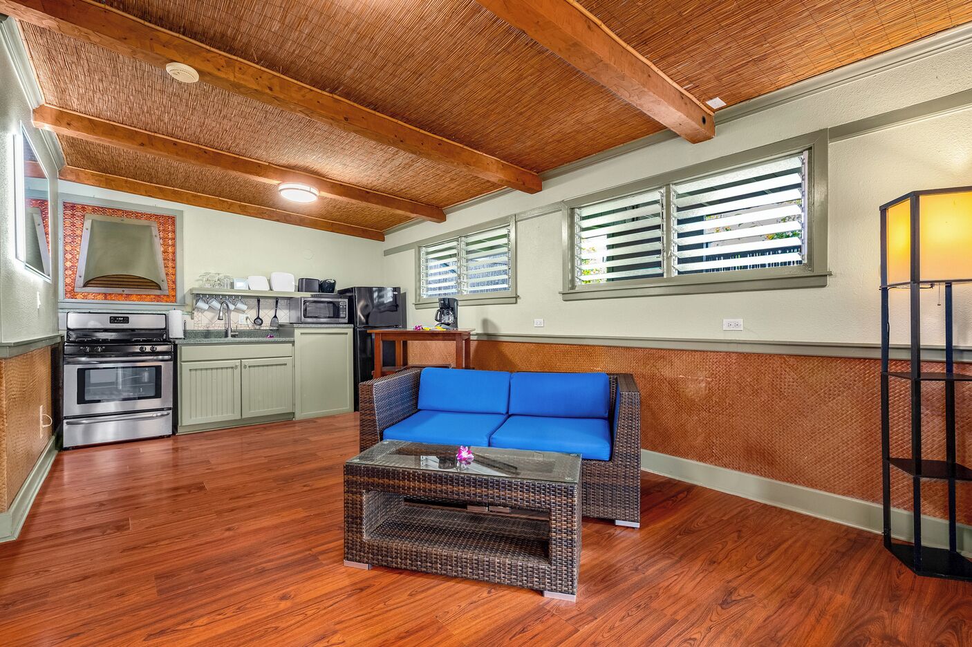 A compact kitchen with a built-in oven, microwave, and coffee maker. A blue cushioned bench is positioned in the center, and a wicker coffee table is in front of it.