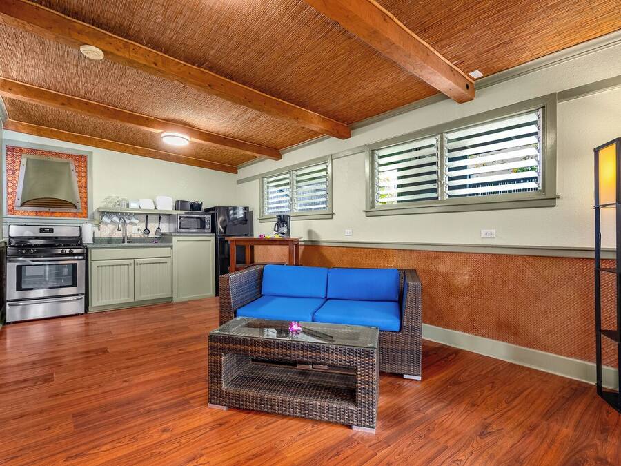 A compact kitchen with a built-in oven, microwave, and coffee maker. A blue cushioned bench is positioned in the center, and a wicker coffee table is in front of it.