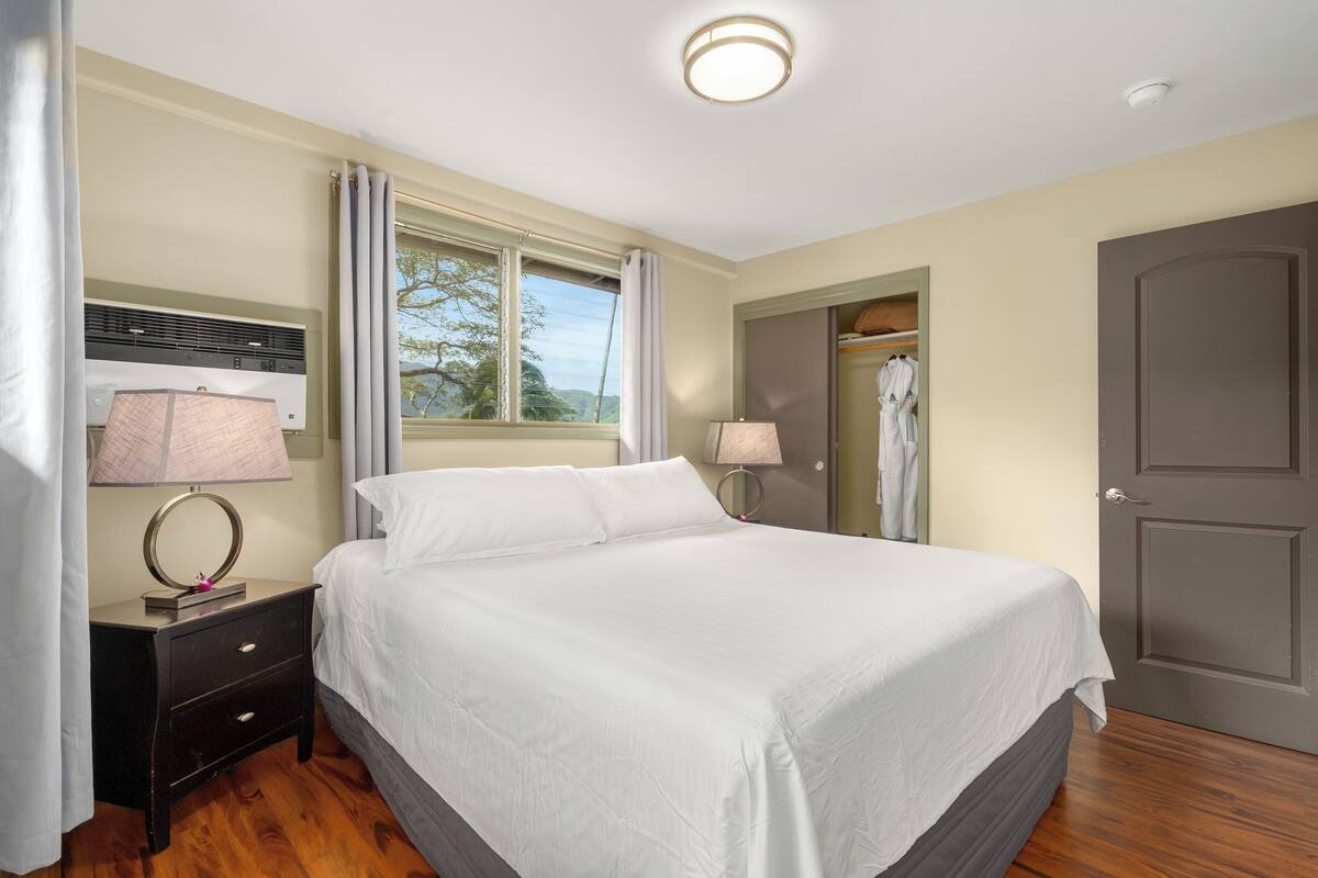 A neatly arranged bedroom with a large bed, two bedside lamps, a nightstand, a window with curtains, and a wardrobe.