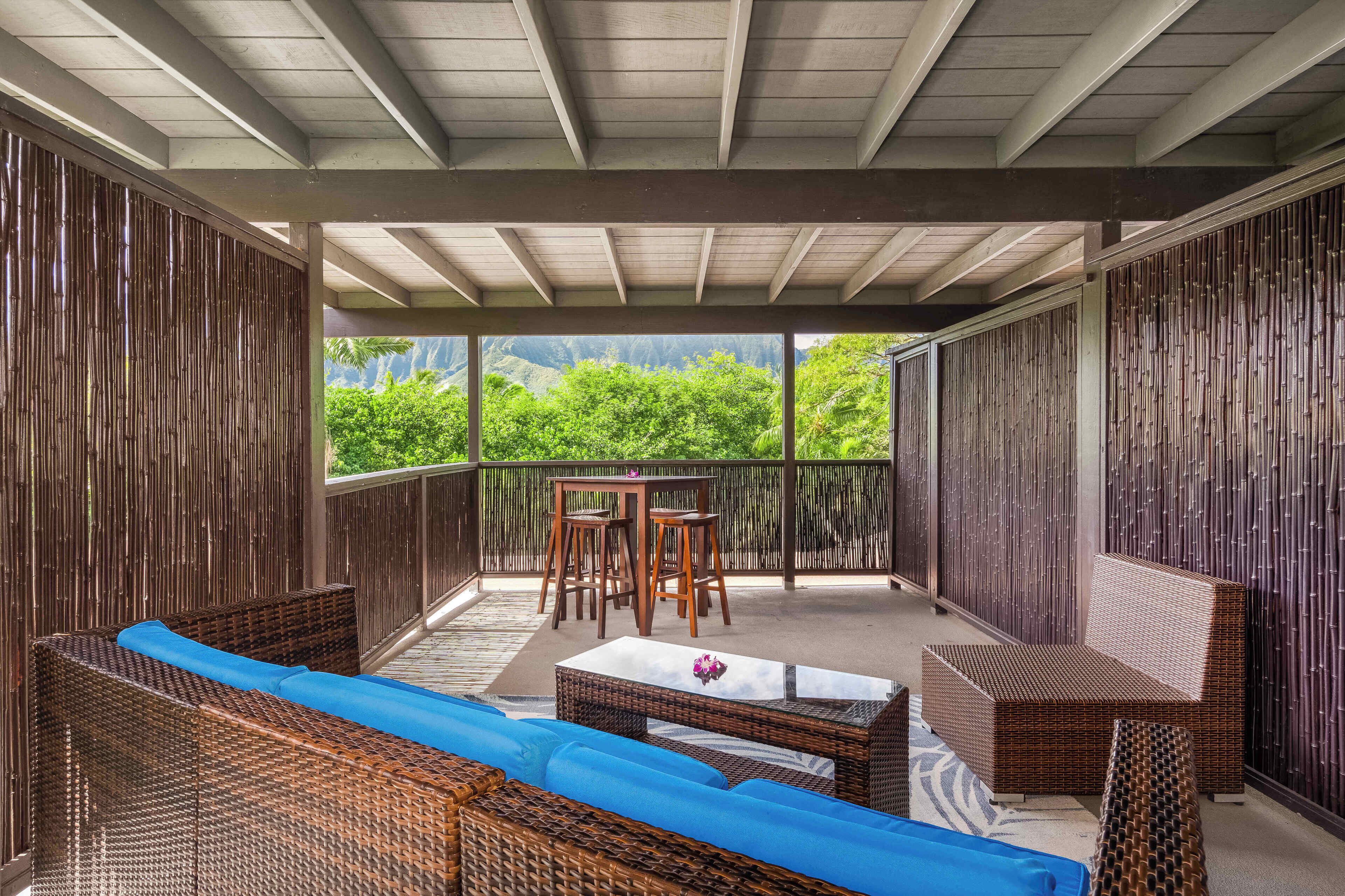A covered outdoor seating area with wicker furniture and a table, overlooking lush greenery.
